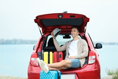 Happy man sitting in car trunk with suitcases on riverside
