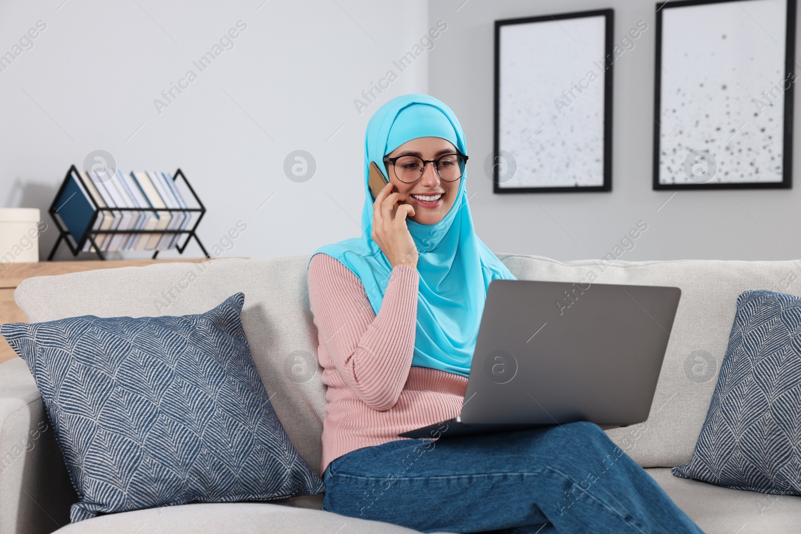 Photo of Muslim woman talking on smartphone near laptop at couch in room