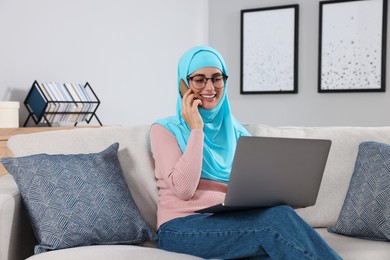 Muslim woman talking on smartphone near laptop at couch in room