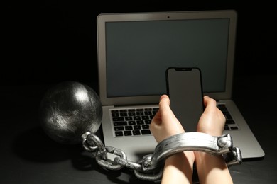 Photo of Woman shackled with ball and chain holding smartphone near laptop on dark background, closeup. Internet addiction