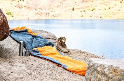 Sleeping bag, thermos and boots on cliff near lake. Space for text