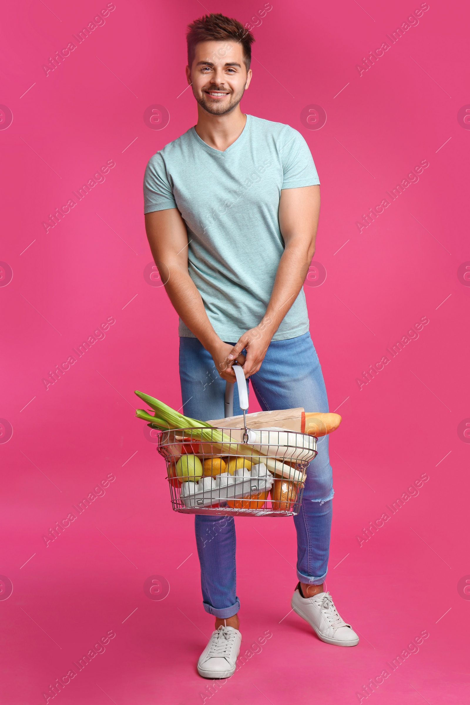 Photo of Young man with shopping basket full of products on pink background