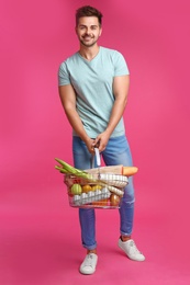 Young man with shopping basket full of products on pink background