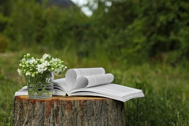Photo of Open book and glass with flowers on tree stump outdoors. Space for text