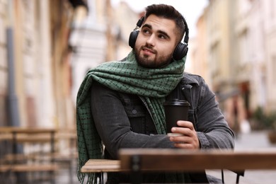 Photo of Smiling man in warm scarf with paper cup listening to music in outdoor cafe