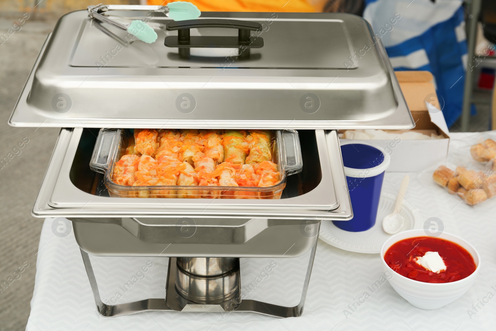 Photo of Volunteer food distribution. Metal warmer with tasty stuffed cabbage and bowl of borsch on white table outdoors