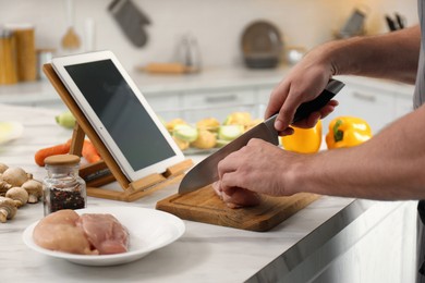 Photo of Man cutting chicken fillet while watching online cooking course via tablet in kitchen, closeup