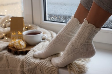 Photo of Woman in warm socks relaxing on window sill at home, closeup