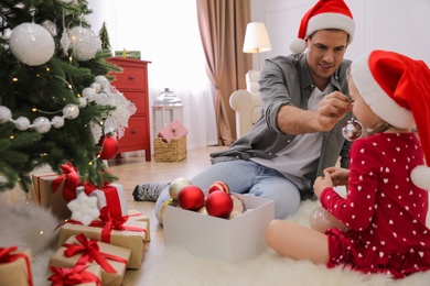 Father with his cute daughter in Santa hats having fun while decorating Christmas tree at home
