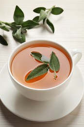 Photo of Cup of aromatic sage tea with fresh leaves on white wooden table