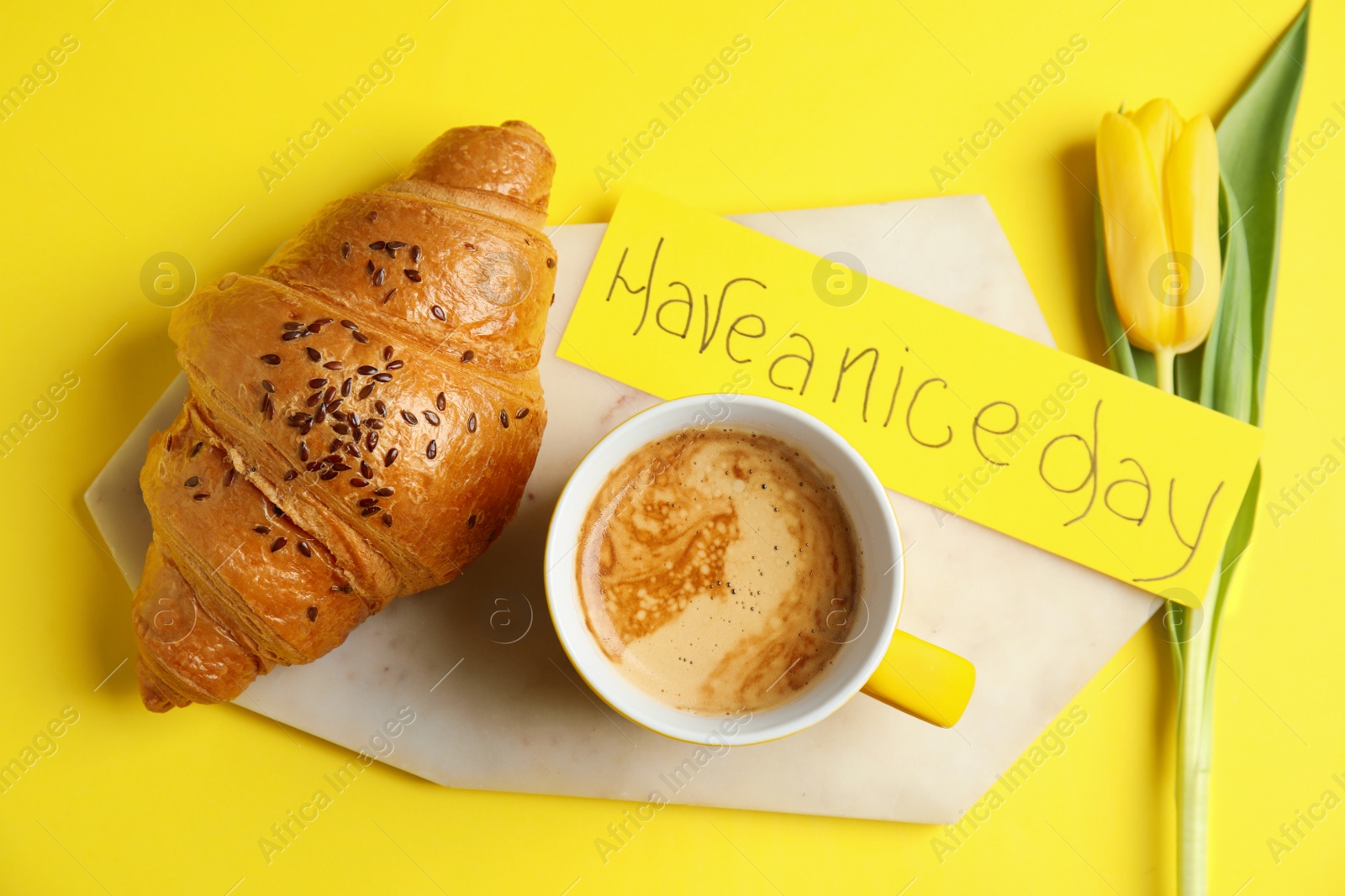 Photo of Delicious morning coffee, croissant, tulip and card with HAVE A NICE DAY wish on yellow background, flat lay