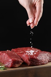 Photo of Woman salting fresh raw beef steak at table against black background, closeup