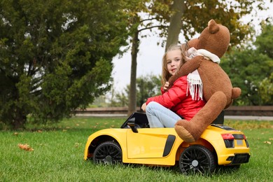 Cute little girl with toy bear driving children's car in park. Space for text