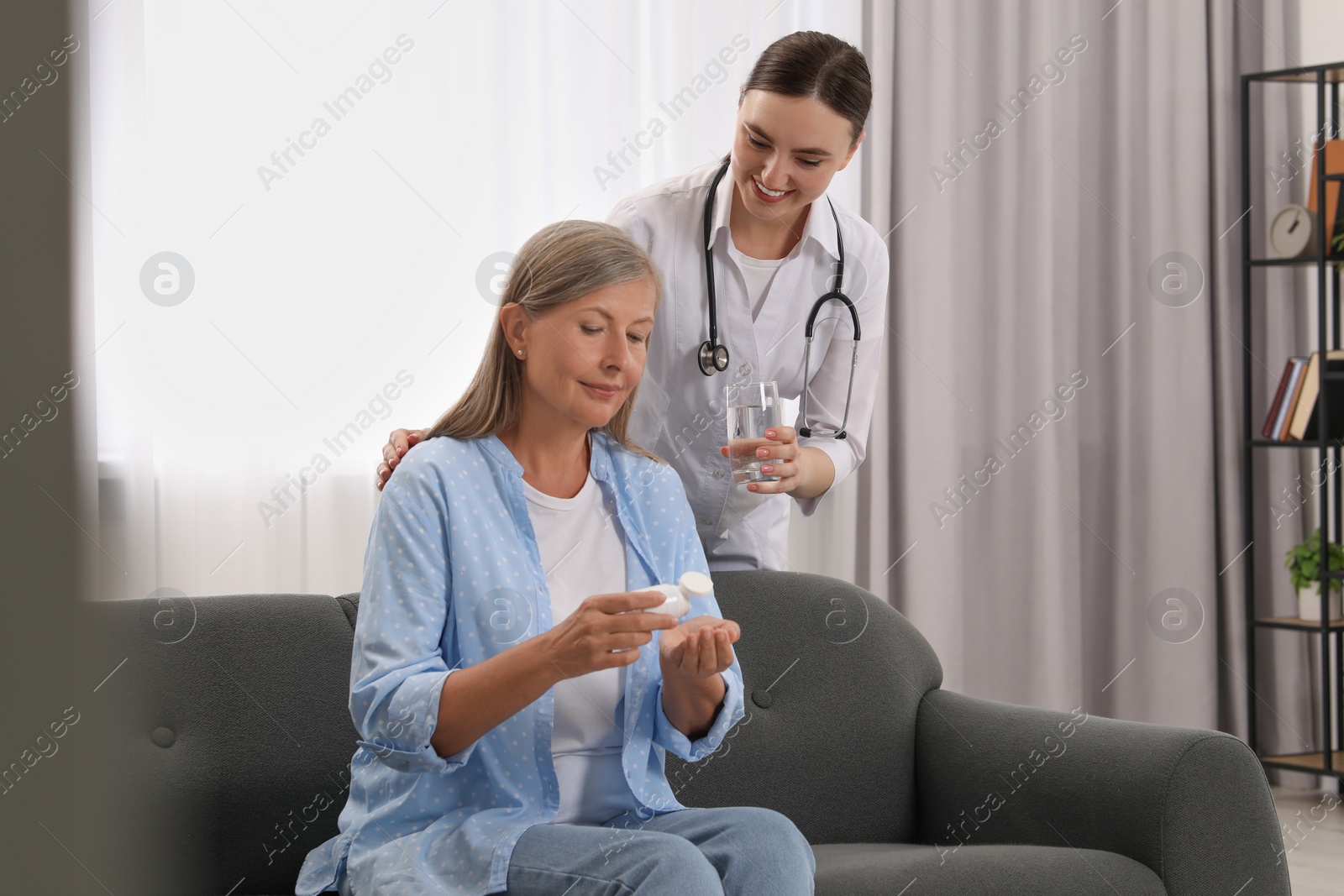 Photo of Young healthcare worker giving glass of water to senior woman with pills indoors