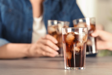 Glass of tasty refreshing cola with ice cubes and blurred people on background