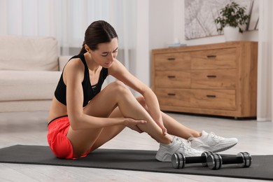 Young woman suffering from leg pain on exercise mat in room