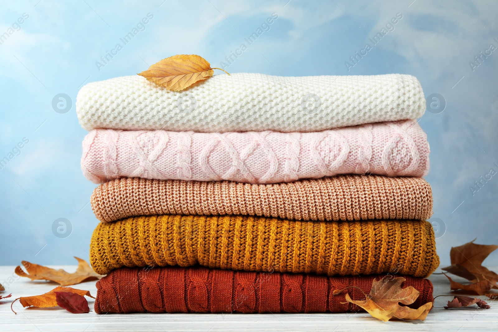 Photo of Stack of warm clothes and autumn leaves on white wooden table against light blue background