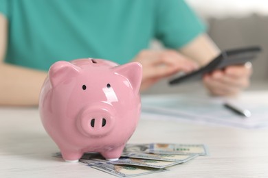 Photo of Financial savings. Woman using calculator at white wooden table indoors, focus on piggy bank and dollar banknotes