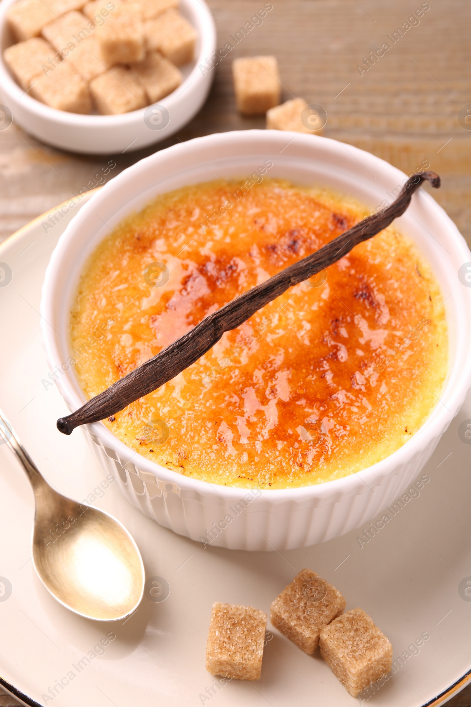 Photo of Delicious creme brulee in bowl, vanilla pod, spoon and sugar cubes on table, closeup