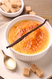 Photo of Delicious creme brulee in bowl, vanilla pod, spoon and sugar cubes on table, closeup