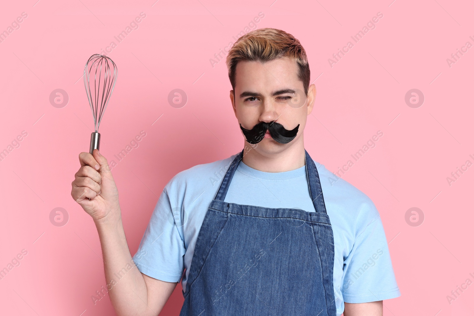 Photo of Portrait of happy confectioner with funny artificial moustache holding whisk on pink background