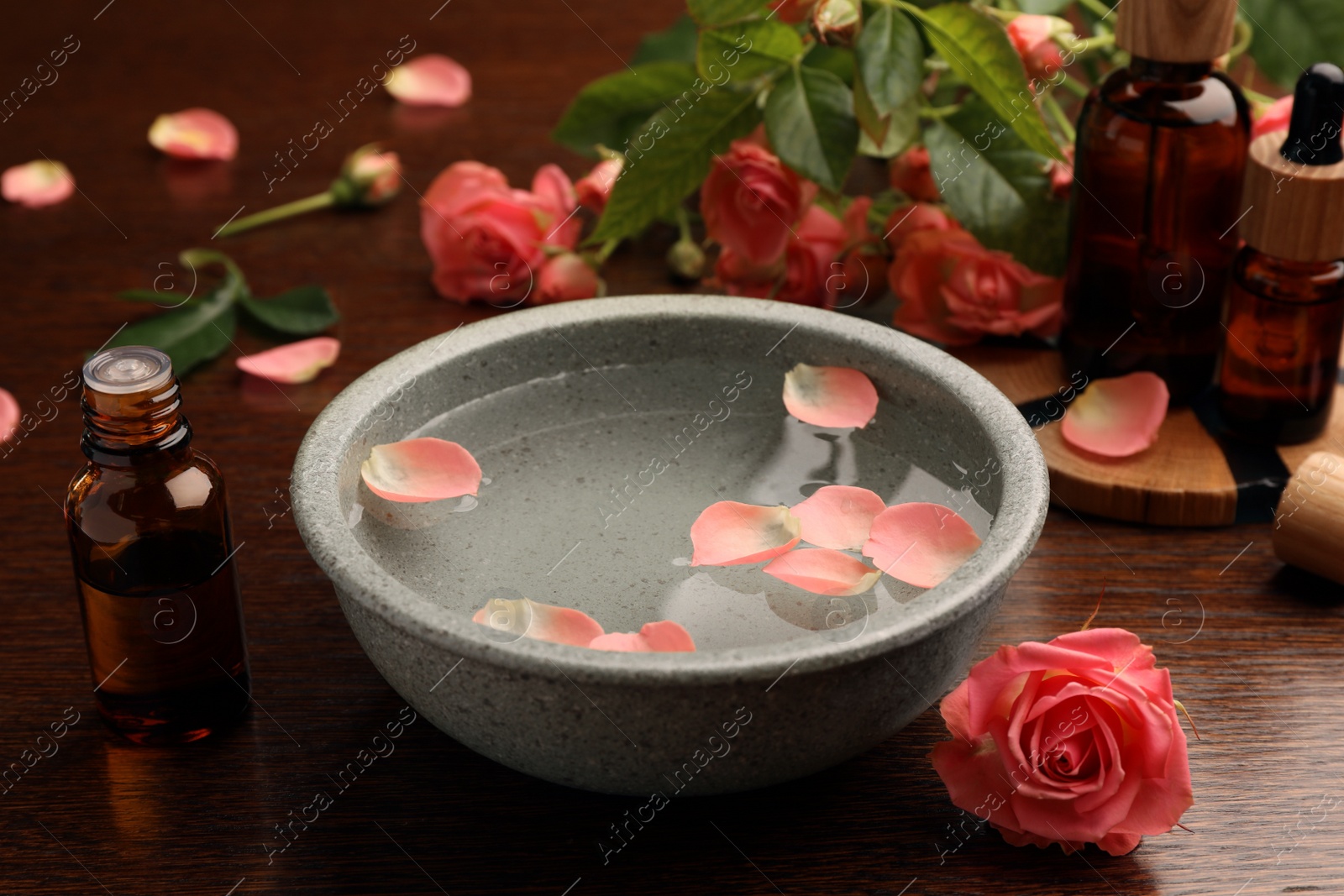 Photo of Bowl of water, bottles with essential oil and beautiful rose petals on wooden table. Aromatherapy treatment