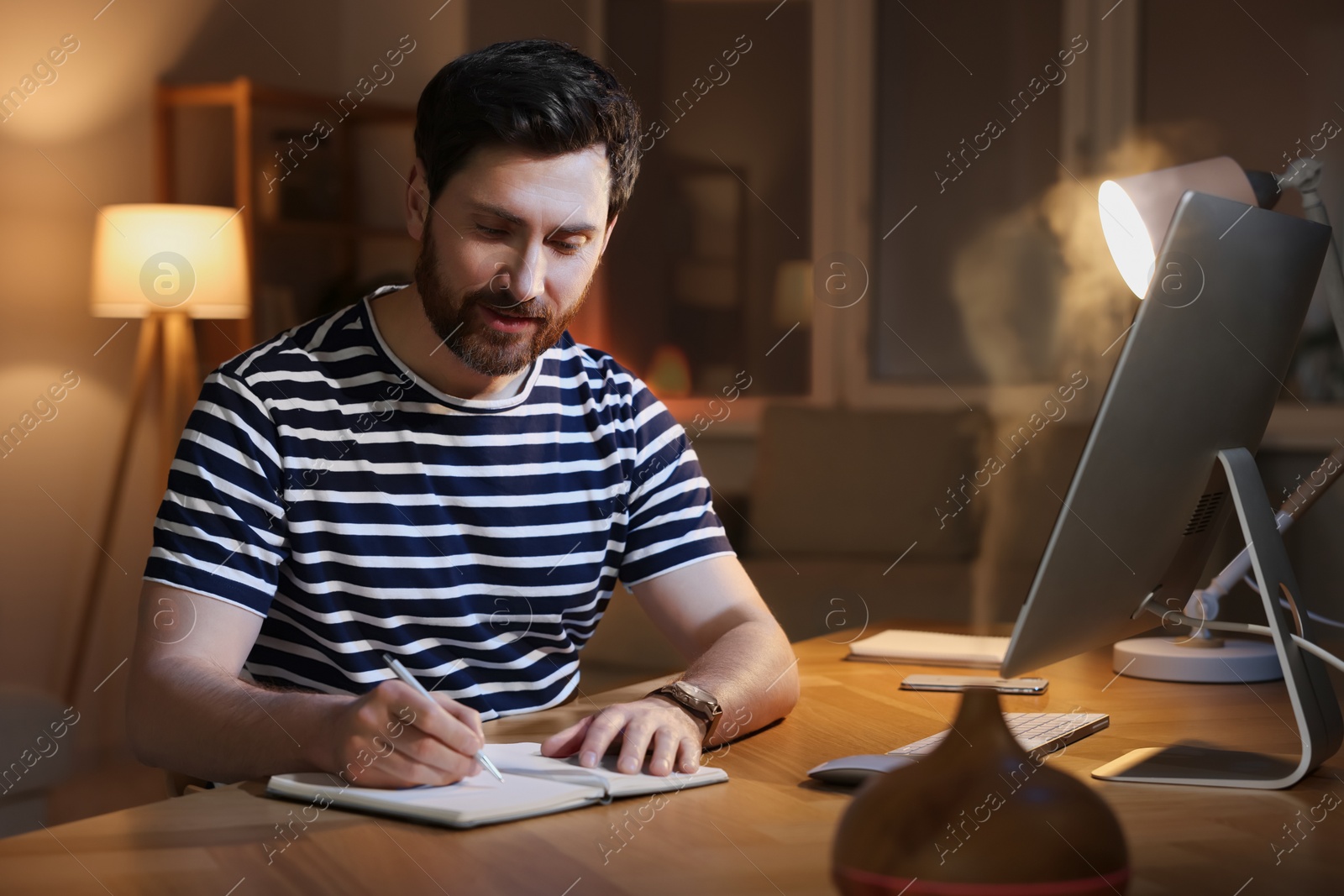 Photo of Home workplace. Man taking notes while working with computer at wooden desk in room at night