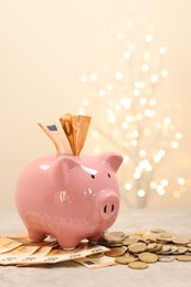 Piggy bank with euro banknotes and coins on grey table against blurred lights, space for text