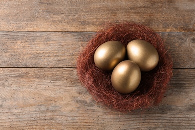 Golden eggs in nest on wooden background, top view with space for text