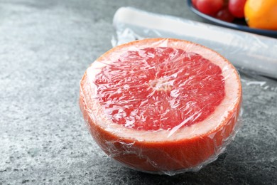 Photo of Half of grapefruit with plastic food wrap on grey table, closeup
