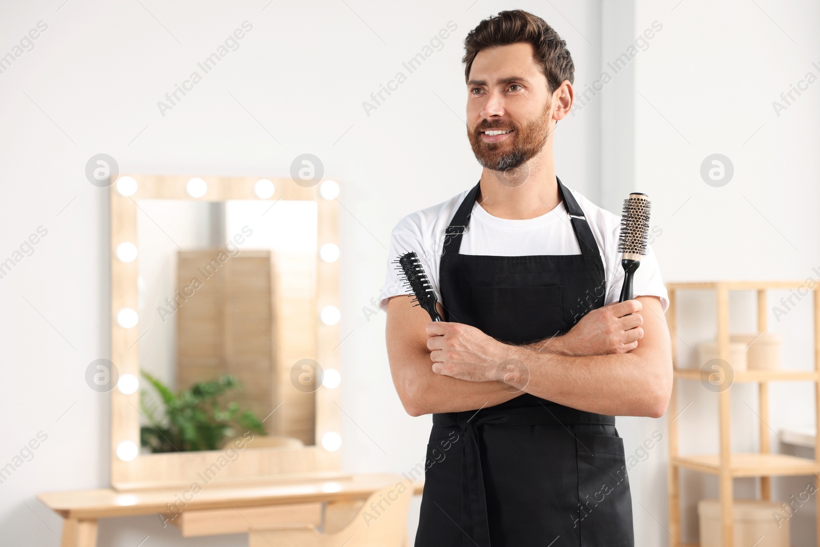 Photo of Smiling hairdresser in apron holding brushes near vanity mirror in salon, space for text