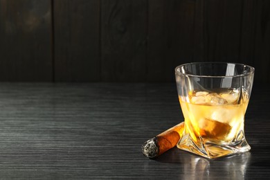Photo of Glass of whiskey with ice cubes and smoldering cigar on black wooden table. Space for text