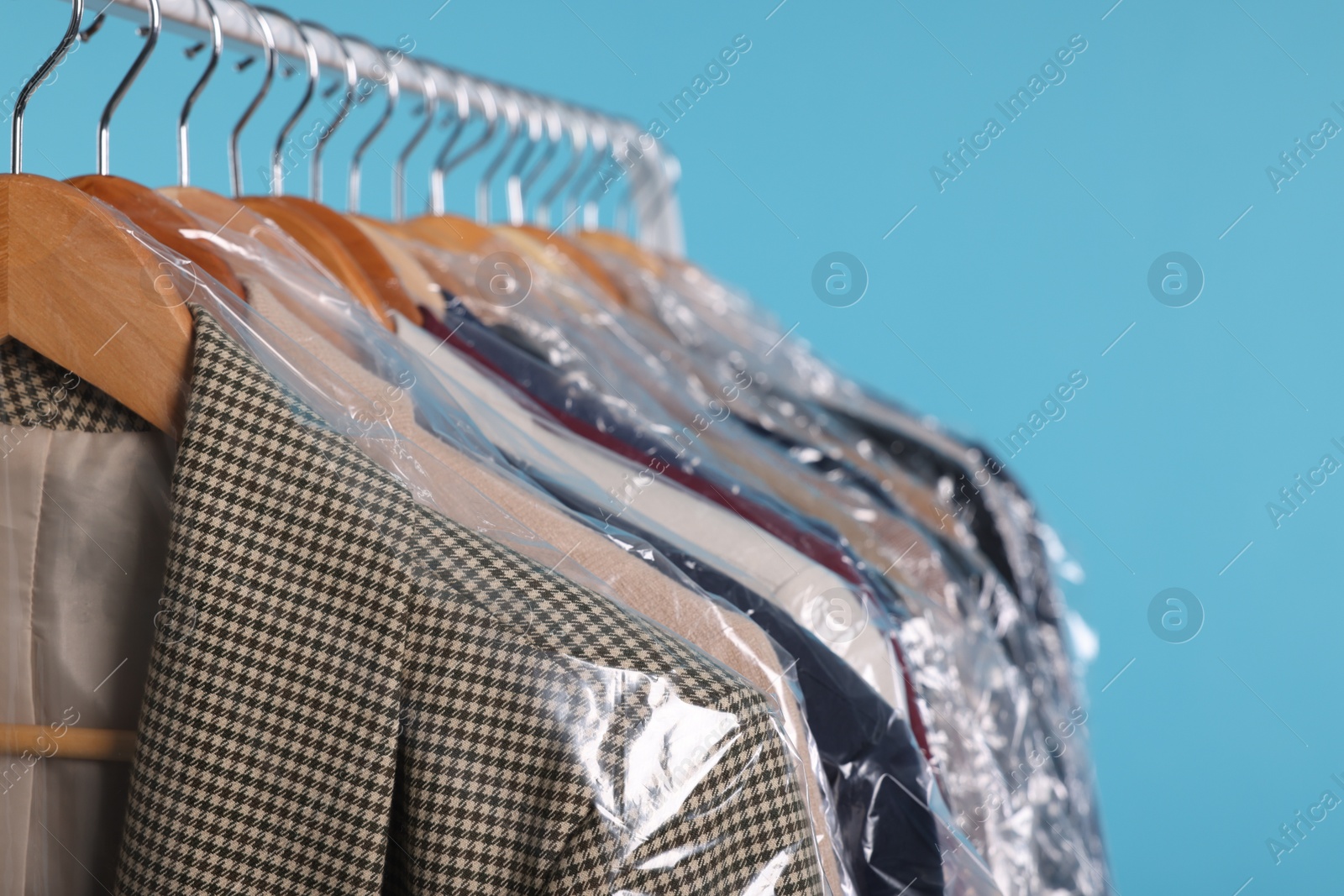 Photo of Dry-cleaning service. Many different clothes in plastic bags hanging on rack against light blue background, closeup