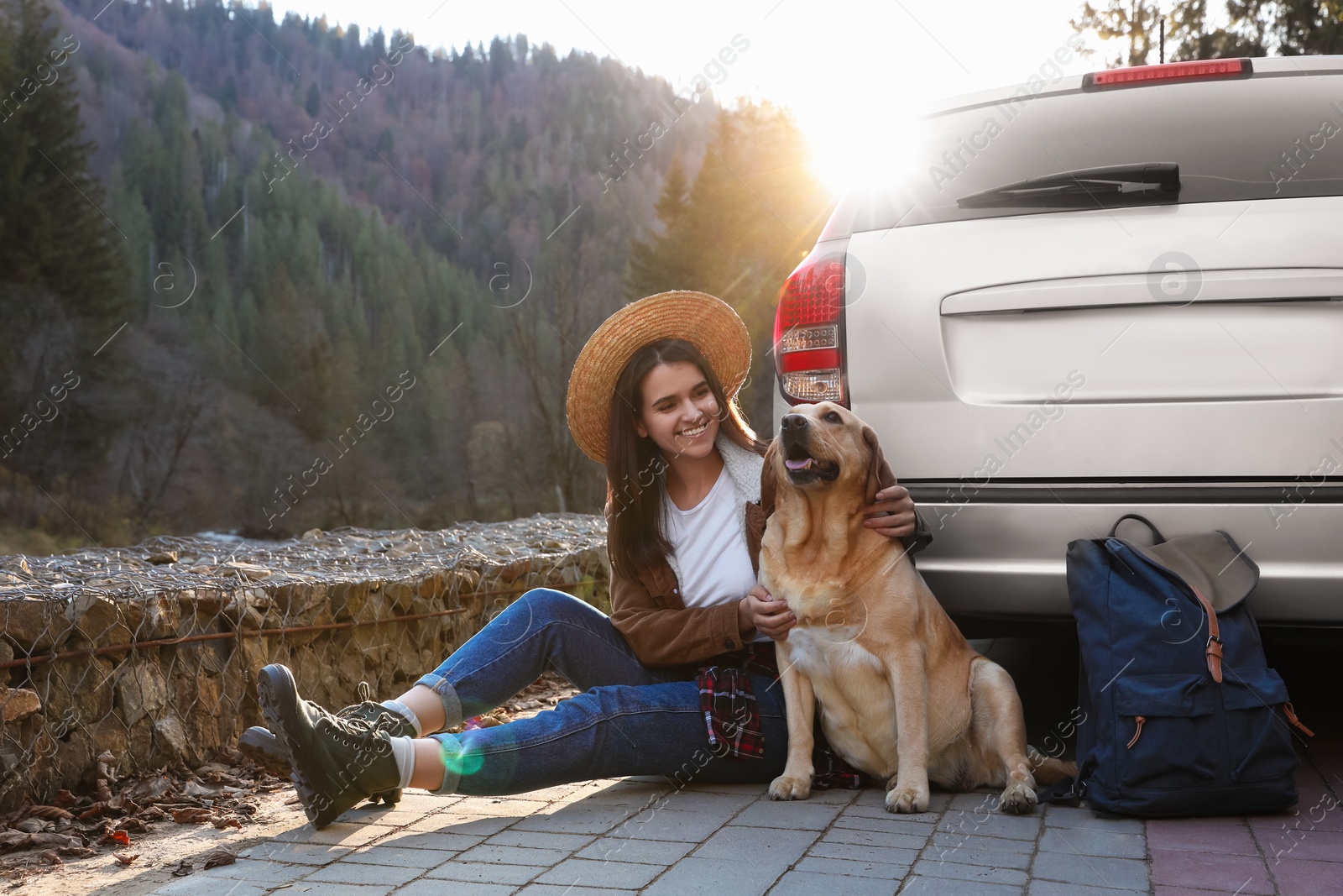 Photo of Happy woman and adorable dog sitting near car in mountains. Traveling with pet