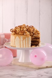 Photo of Caramel drip cake decorated with popcorn and pretzels near balloons and gift on white marble table