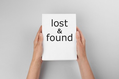 Woman holding book with phrase Lost and Found on light grey background, closeup