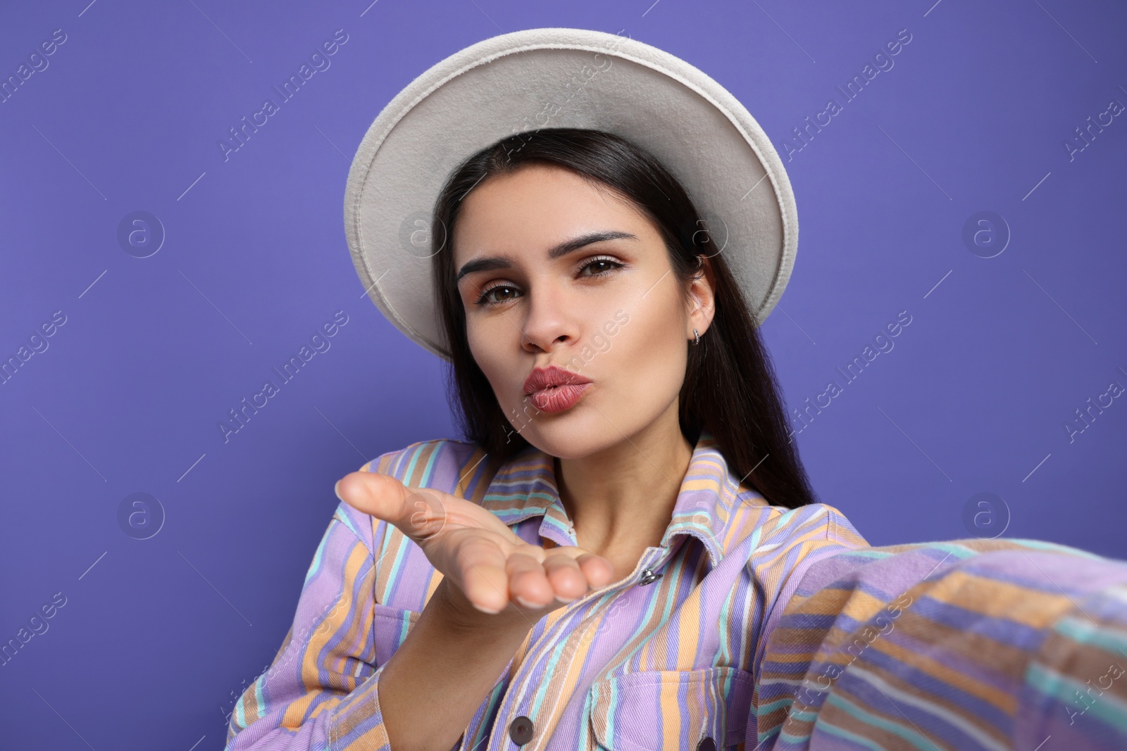 Photo of Beautiful young woman with stylish hat taking selfie while blowing kiss on purple background, closeup