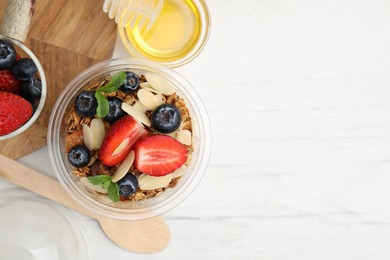 Photo of Tasty granola with berries, almond flakes and mint in plastic cup on white wooden table, flat lay. Space for text