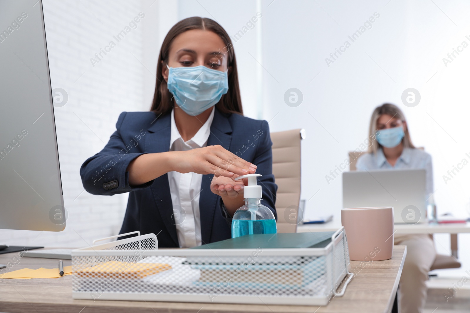 Photo of Office employee in mask applying hand sanitizer at workplace