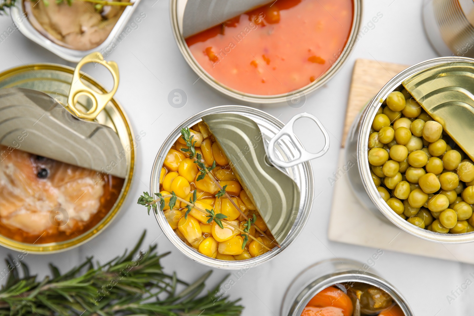 Photo of Open tin cans with different products and rosemary on white table, flat lay