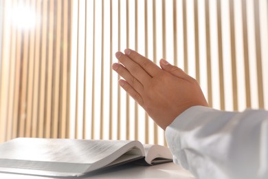 Woman holding hands clasped while praying over Bible at white table indoors, closeup