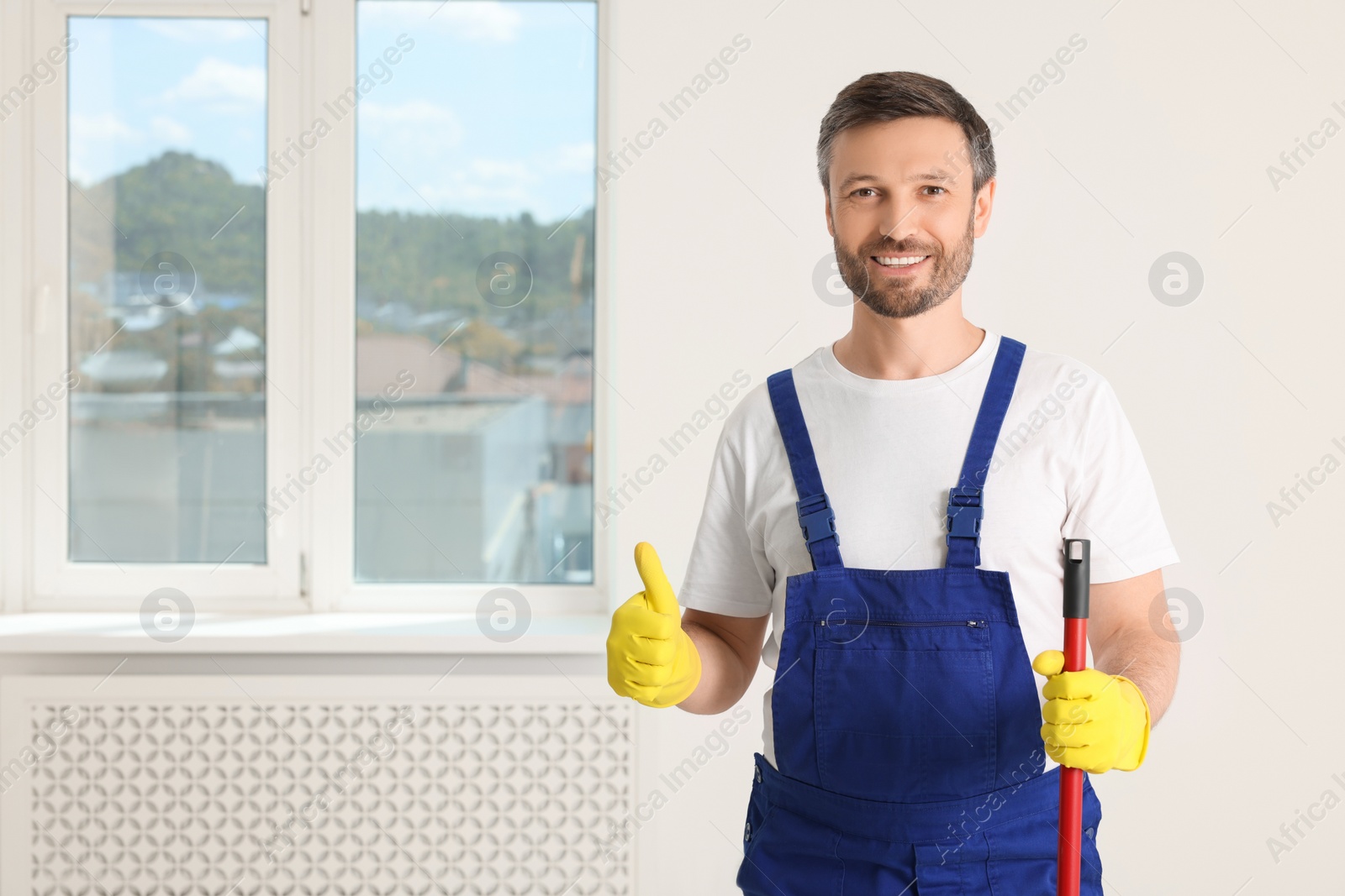 Photo of Man in uniform with mop showing thumb up indoors. Space for text