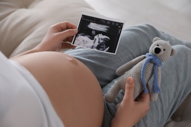 Photo of Young pregnant woman with ultrasound picture of baby and toy bear indoors, closeup