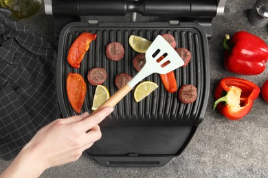 Photo of Woman cooking meat balls with bell peppers and lemon on electric grill at grey textured table, top view