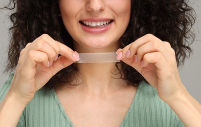 Photo of Young woman holding teeth whitening strip on grey background, closeup
