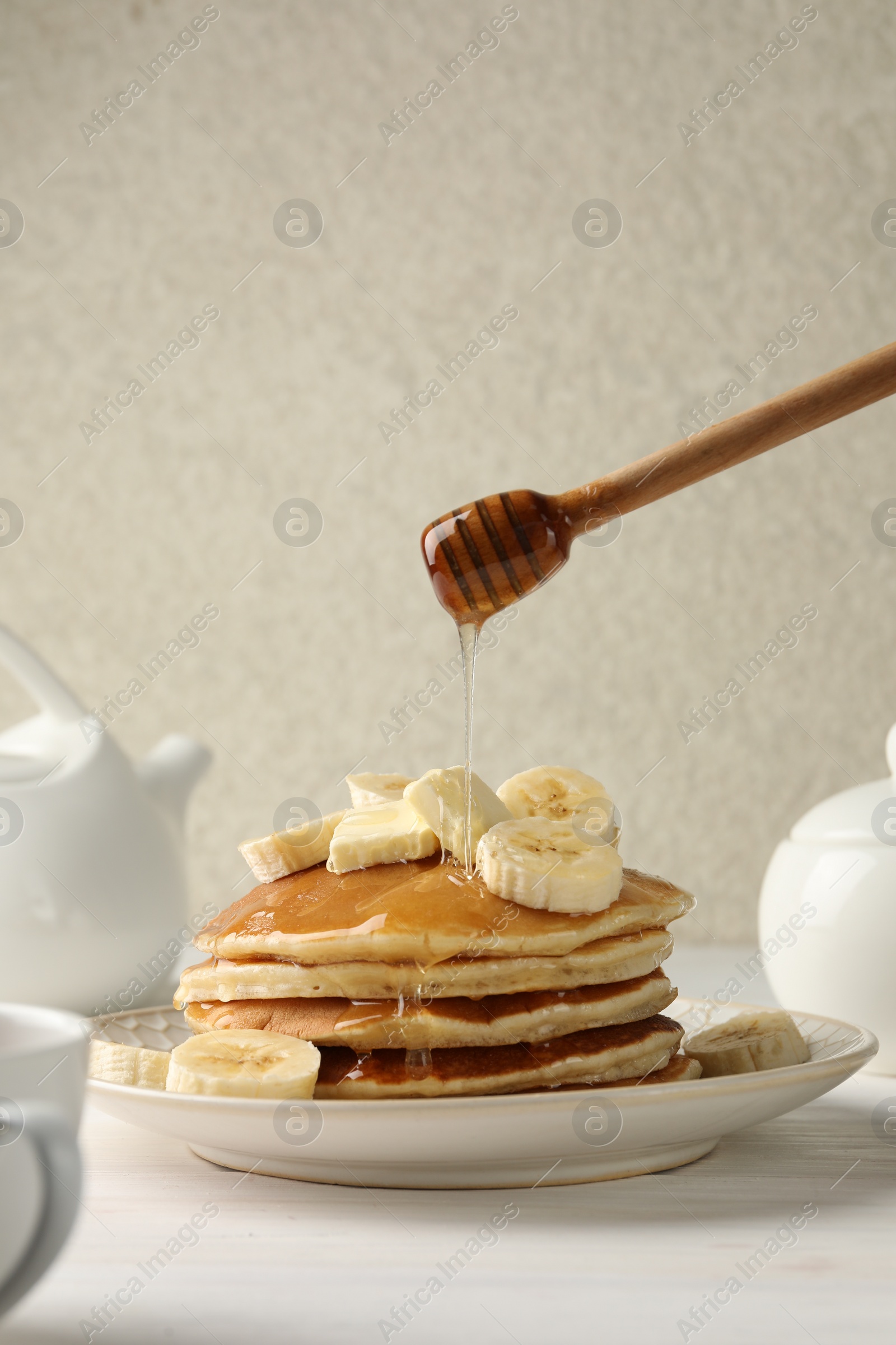 Photo of Pouring honey from dipper onto delicious pancakes with bananas and butter at white table