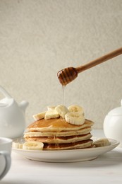 Photo of Pouring honey from dipper onto delicious pancakes with bananas and butter at white table