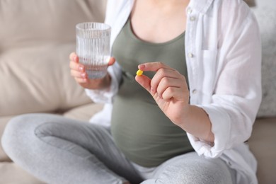 Photo of Pregnant woman holding pill and glass with water on sofa, closeup
