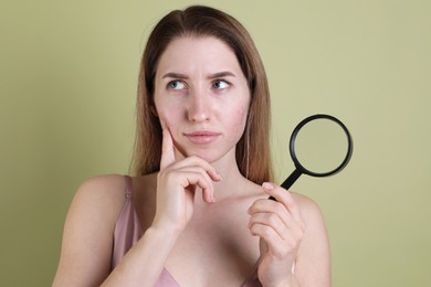 Photo of Thoughtful young woman with acne problem holding magnifying glass on olive background