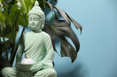 Photo of Buddhism religion. Decorative Buddha statue with burning candle and monstera against light blue wall, space for text